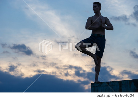 Fit young man practices sun salutation yoga on the edge of cliff at sunset 63604604