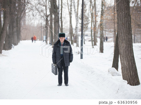 An old man with suitcase standing outdoors in winter park while snowfall An old man with suitcase standing outdoors in winter park while snowfall 63630956