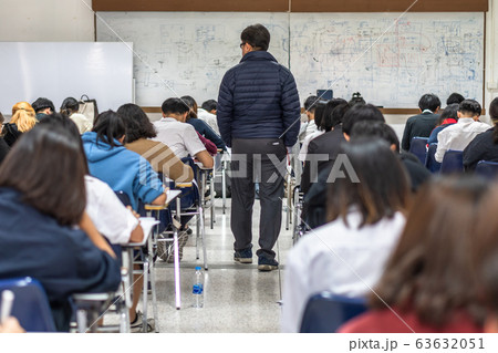 School exam room with teacher (invigilator) monitoring students taking educational examination in classroom for admission test School exam room with teacher (invigilator) monitoring students taking educational examination in classroom for admission test 63632051