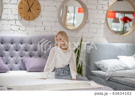 Blonde woman with jewelry on neck sitting on the bed. 63634102