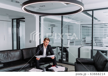 a young stylish businessman is sitting on a sofa in a modern office and holding important documents in his hands 63668214