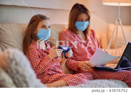 Woman in pajamas with notebook and papers working from home wearing protective mask while her kid, daughter playing computer console games 63676702