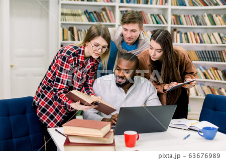 Group of smart modern confident multiethnic friends, preparing together for exams in the college library. African boy is using laptop, blond pretty girl showing him the book 63676989