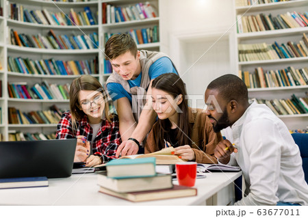Four positive smiling multiethnic boys and girls sitting at the table in modern college library and jointly doing home task or searching information for learning subject Four positive smiling multiethnic boys and girls sitting at the table in modern college library and jointly doing home task or searching information for learning subject 63677011