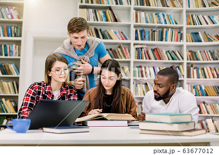 Group of confident smart attractive young mixed race friends, students or coworkers, which working together in the library, preparing for exams, practical class or new project, using laptop and books 63677012