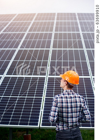 Engineer in an orange helmet stands with his back to camera against the background of large plantation solar batteries Engineer in an orange helmet stands with his back to camera against the background of large plantation solar batteries 63699910