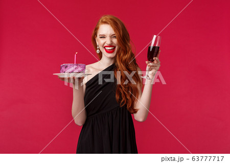 Portrait of excited laughing gorgeous redhead woman having fun at b-day party, holding glass of wine and birthday cake with lit candle, making wish, celebrating over red background Portrait of excited laughing gorgeous redhead woman having fun at b-day party, holding glass of wine and birthday cake with lit candle, making wish, celebrating over red background 63777717