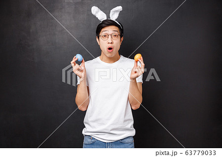 Holidays, party and Easter concept. Amazed young asian guy in rabbit ears holding painted eggs as learning girlfriend traditions, open mouth fascinated get to know new culture, black background 63779033