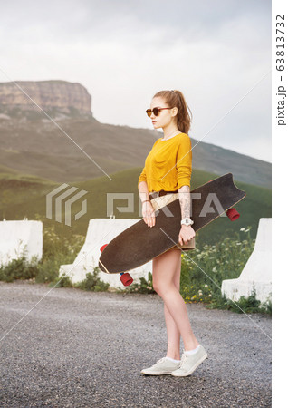 Beautiful and fashionable young woman in sunglasses and with a tattoo poses with a skateboard or longboard against the sunset sky 63813732