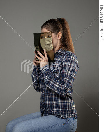 Be safe and read to become someone else - woman covering face with book in face mask while reading on grey background Be safe and read to become someone else - woman covering face with book in face mask while reading on grey background 63846981