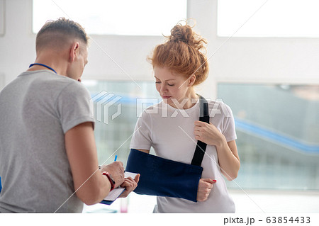 Male physiotherapist writing instructions, female patient standing in front of him in arm sling 63854433