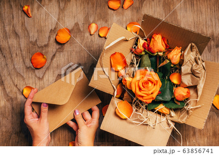 Woman's hands open craft paper envelope next to open box with orange roses in it, close up. Woman's hands open craft paper envelope next to open box with orange roses in it, close up. 63856741