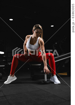 Attractive shape young sporty focused fitness girl with ponytail doing biceps exercises while sitting on the bench and raising dumbell in the gym at night. Attractive shape young sporty focused fitness girl with ponytail doing biceps exercises while sitting on the bench and raising dumbell in the gym at night. 63863005