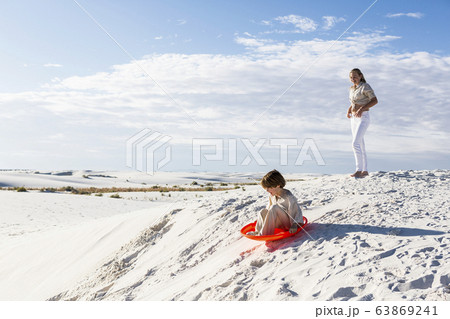 Children playing in sand dunes landscape, one on an orange sled. 63869241
