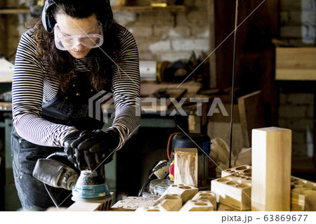 Woman with long brown hair wearing dungarees, safety glasses and ear protectors standing in wood workshop, using sander. 63869677