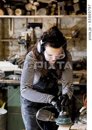 Woman with long brown hair wearing dungarees, safety glasses and ear protectors standing in wood workshop, using sander. 63869797