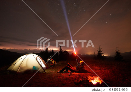 Woman resting at night camping near campfire, tourist tent, bicycle under evening sky full of stars 63891466