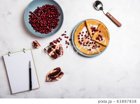 Grains of peeled pomegranate lie in a plate. Pancakes with pomegranate and condensed milk lie on a plate. View from above. Close-up. A book for writing recipes lies on the table. Copy space for text. 63897838