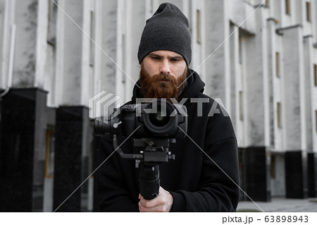 Bearded Professional videographer in black hoodie holding professional camera on 3-axis gimbal stabilizer. Filmmaker making a great video with a professional cinema camera. Cinematographer. 63898943