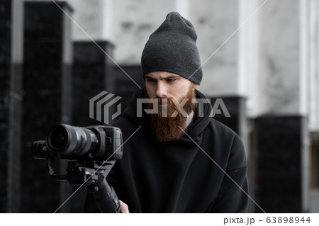 Bearded Professional videographer in black hoodie holding professional camera on 3-axis gimbal stabilizer. Filmmaker making a great video with a professional cinema camera. Cinematographer. 63898944