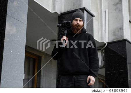 Bearded Professional videographer in black hoodie holding professional camera on 3-axis gimbal stabilizer. Filmmaker making a great video with a professional cinema camera. Cinematographer. 63898945