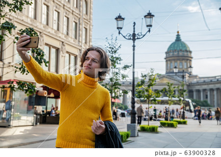 The handsome men does selfie, Look from outside, he dressed in a yellow sweater, a black raincoat or jacket is his hands, Bolshaya Konyushennaya street and Kazan Cathedral on background, sunny day 63900328