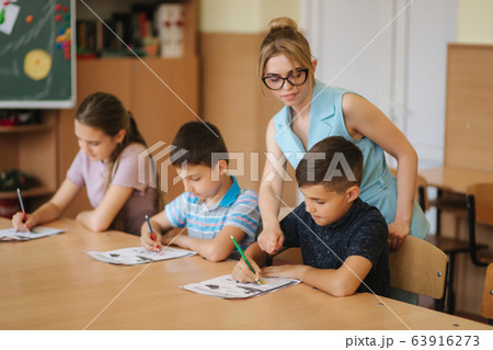 Teacher helping school kids writing test in classroom. education, elementary school, learning and people concept Teacher helping school kids writing test in classroom. education, elementary school, learning and people concept 63916273