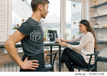 Interior of coffee shop, young woman talking with cafe worker with cup of coffee at table 63923106