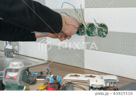 Renovation and construction in kitchen, close-up of electricians hand installing outlet on wall with ceramic tiles using professional tools 63925510