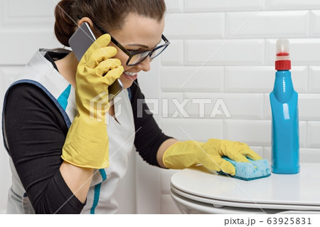Adult positive woman doing cleaning with detergents. Woman in glasses, professional uniform in bathroom, toilets, woman laughing and talking on phone 63925831