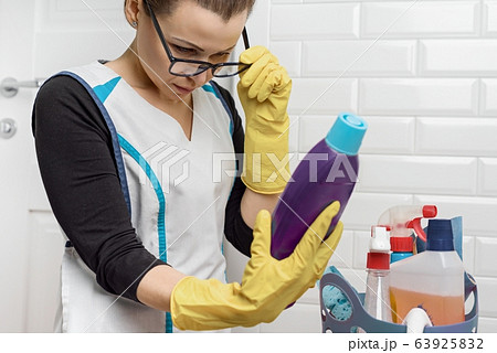 Adult woman doing cleaning with detergents. Woman in glasses, professional uniform in bathroom, toilets room, reading instructions on cleaning bottle Adult woman doing cleaning with detergents. Woman in glasses, professional uniform in bathroom, toilets room, reading instructions on cleaning bottle 63925832