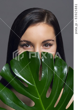 Studio beauty portrait of young brunette with natural make-up perfect skin with green exotic leaf on gray background. Concept of natural cosmetics, face and body care products. Studio beauty portrait of young brunette with natural make-up perfect skin with green exotic leaf on gray background. Concept of natural cosmetics, face and body care products. 63925881