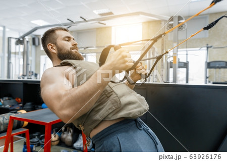 Muscular bearded man dressed in military weighted armored vest doing exercises using straps systems in the gym. Sport, training, bodybuilding and healthy lifestyle concept. Muscular bearded man dressed in military weighted armored vest doing exercises using straps systems in the gym. Sport, training, bodybuilding and healthy lifestyle concept. 63926176