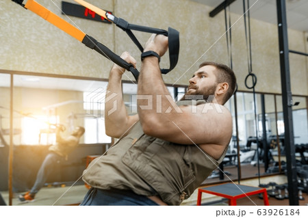 Muscular bearded man dressed in military weighted armored vest doing exercises using straps systems in the gym. Sport, training, bodybuilding and healthy lifestyle concept. Muscular bearded man dressed in military weighted armored vest doing exercises using straps systems in the gym. Sport, training, bodybuilding and healthy lifestyle concept. 63926184