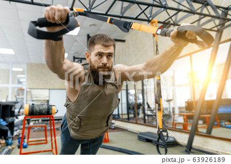Muscular bearded man dressed in military weighted armored vest doing exercises using straps systems in the gym. Sport, training, bodybuilding and healthy lifestyle concept. Muscular bearded man dressed in military weighted armored vest doing exercises using straps systems in the gym. Sport, training, bodybuilding and healthy lifestyle concept. 63926189
