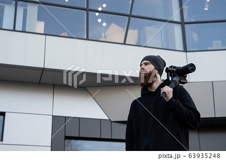 Bearded Professional videographer in black hoodie holding professional camera on 3-axis gimbal stabilizer. Filmmaker making a great video with a professional cinema camera. Cinematographer. 63935248