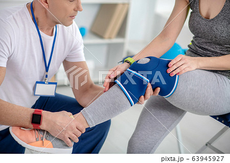 Male physiotherapist examining knee of female patient sitting on couch Male physiotherapist examining knee of female patient sitting on couch 63945927