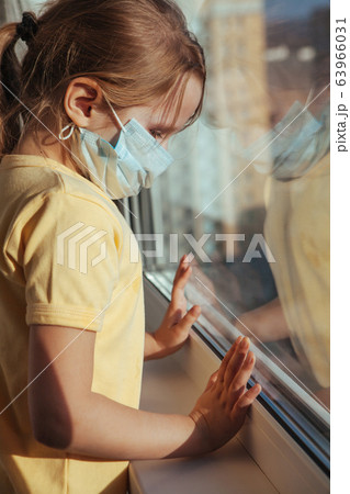 A little girl in a medical mask looks out the window. Coronavirus quarantine. A little girl in a medical mask looks out the window. Coronavirus quarantine. 63966031