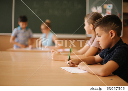 Education, elementary school. Learning and people concept - group of school kids with pens and notebooks writing test in classroom Education, elementary school. Learning and people concept - group of school kids with pens and notebooks writing test in classroom 63967506