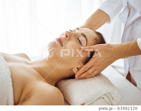Beautiful brunette woman enjoying facial massage with closed eyes. Relaxing treatment in medicine and spa center concepts Beautiful brunette woman enjoying facial massage with closed eyes. Relaxing treatment in medicine and spa center concepts 63982120