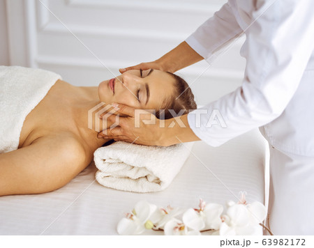 Beautiful brunette woman enjoying facial massage with closed eyes. Relaxing treatment in medicine and spa center concepts Beautiful brunette woman enjoying facial massage with closed eyes. Relaxing treatment in medicine and spa center concepts 63982172