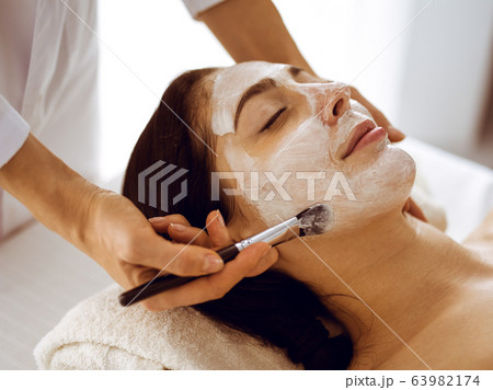 Beautiful brunette woman enjoying applying cosmetic mask with closed eyes. Relaxing treatment in medicine and spa center concepts Beautiful brunette woman enjoying applying cosmetic mask with closed eyes. Relaxing treatment in medicine and spa center concepts 63982174