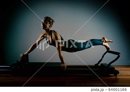 Young girl doing pilates exercises with a reformer bed. Beautiful slim fitness trainer on reformer gray background, low key, art light. Fitness concept Young girl doing pilates exercises with a reformer bed. Beautiful slim fitness trainer on reformer gray background, low key, art light. Fitness concept 64001166