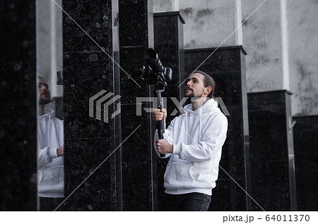 Young Professional videographer holding professional camera on 3-axis gimbal stabilizer. Pro equipment helps to make high quality video without shaking. Cameraman wearing white hoodie making a videos. 64011370