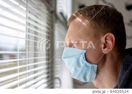 Man in a medical face mask looks out the window through the blinds. Quarantine during an epidemic. 64020524