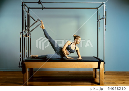 Young girl doing pilates exercises with a reformer bed. Beautiful slim fitness trainer on reformer gray background, low key, art light. Fitness concept 64028210