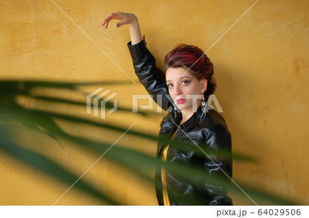 Glamorous lady in punk rock style party clothes with dark pink hair in casual dress, black leather jacket, and long earrings on yellow background 64029506