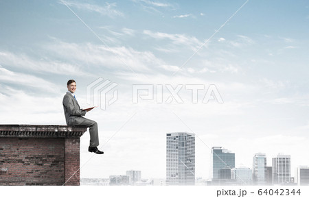 Man on roof edge reading book and cityscape at background Man on roof edge reading book and cityscape at background 64042344