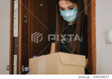 Young woman in maks and gloves take a parcel by her self at home. Package on the floor by the door. Self-qurantine. Coronavirus concept 64059657