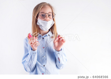 Small pretty girl in a medical mask is holding antiseptic bottle in her hands against light grey background. 64068566